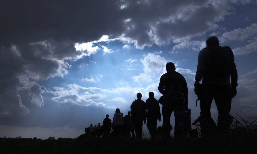 Silhoutte of a line of immigrants fleeing climate change under a dark and threatening sky.