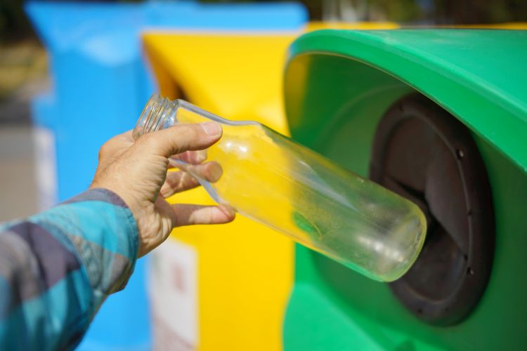 A man's hand placing a glass bottle into a green recycling bin.