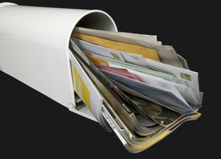 Photo of a white mailbox tuffed wiht junk mails and magazines on dark grey background.