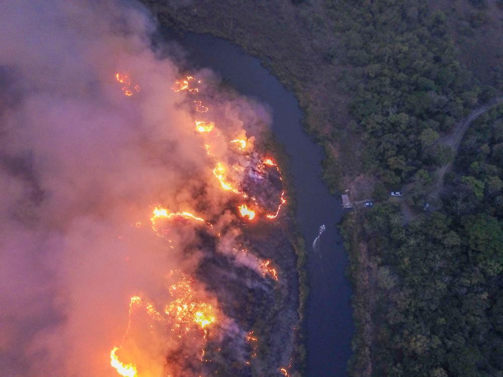 Brilliant red, orange flames burning along a riverbank, with gray smoke rising in columns.