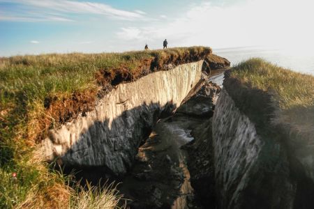 Permafrost damage in the center of grass covered Alaskan cliffs, with two people looking out towards the ocean. The sunken center area shows a white sub- soil color and debris in the bottom.