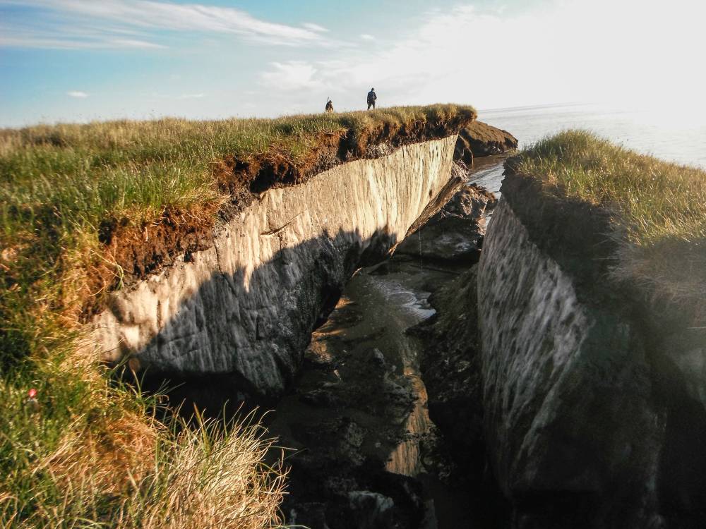 Permafrost damage in the center of grass covered Alaskan cliffs, with two people looking out towards the ocean. The sunken center area shows a white sub- soil color and debris in the bottom.