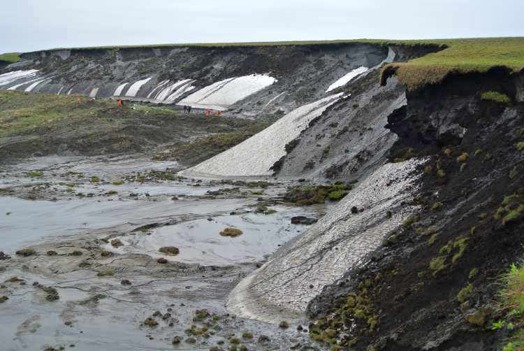 Herschel Island image showing permafrost sunken foreground of soil and water, with dwarfed people standing in it, and approximately 100 feet lower than the dark, black soil and melting ice sidewall, which has green grass on the top of it and a light blue sky overhead.