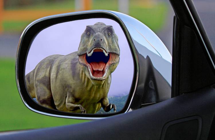 A green T-REX dino looks through the rear mirror of a car. Its red mouth opens with white teeth and tongue, its font limps are small.