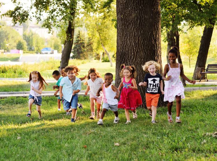 A group of preschoolers running on the grass in the Park. They wear different clothes colors such as white, red, blue, pink , navy, teal and orange. Boys are in short jeans and girls re in dresses and short.